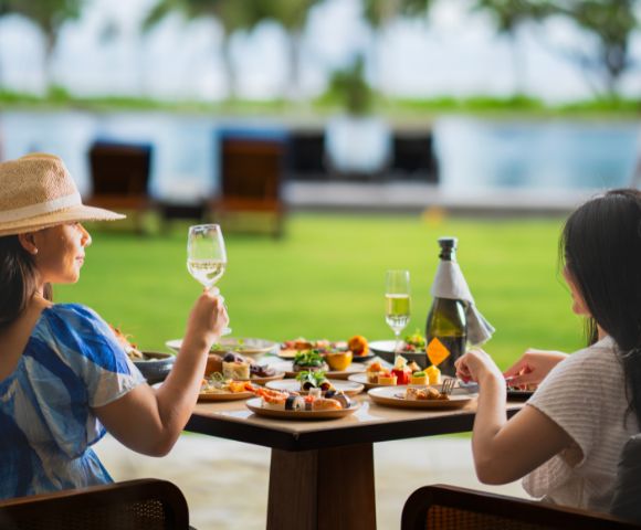 Two women seated at a table outdoors, enjoying a meal with a variety of dishes and wine. They appear relaxed, with a scenic pool and greenery in the background.