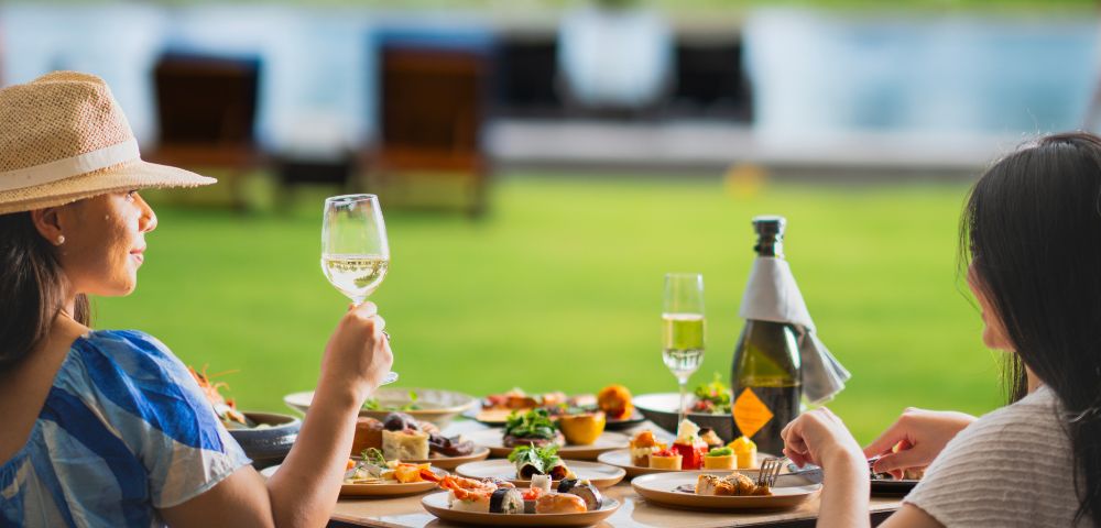 Two women seated at a table outdoors, enjoying a meal with a variety of dishes and wine. They appear relaxed, with a scenic pool and greenery in the background.