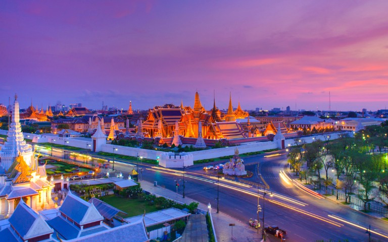 A vibrant view of the Grand Palace in Bangkok at sunset, with illuminated spires against a colorful pink and purple sky. Busy streets and lush green parks add to the serene yet lively ambiance.