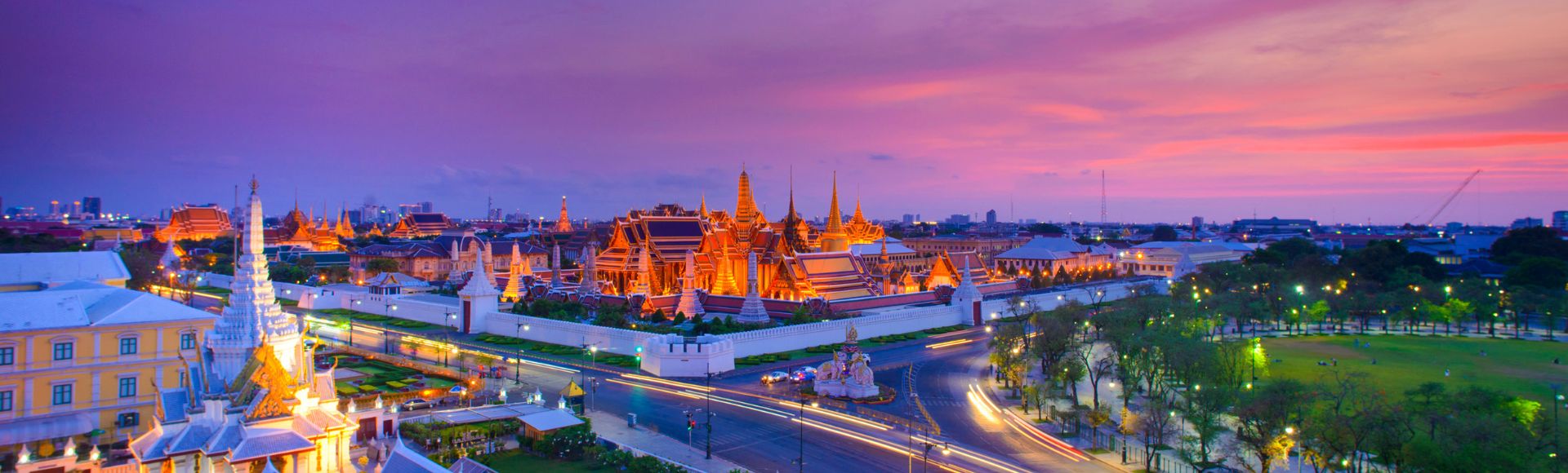 A vibrant view of the Grand Palace in Bangkok at sunset, with illuminated spires against a colorful pink and purple sky. Busy streets and lush green parks add to the serene yet lively ambiance.