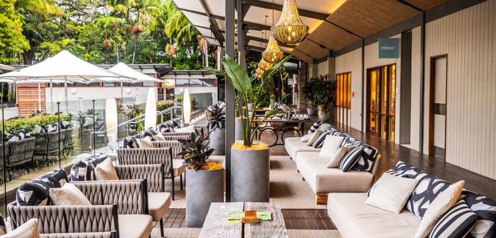 Spacious outdoor patio with plush sofas, patterned cushions, and modern decor. Hanging lights and plants add warmth and elegance, with lush greenery visible.