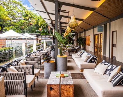 Spacious outdoor patio with plush sofas, patterned cushions, and modern decor. Hanging lights and plants add warmth and elegance, with lush greenery visible.