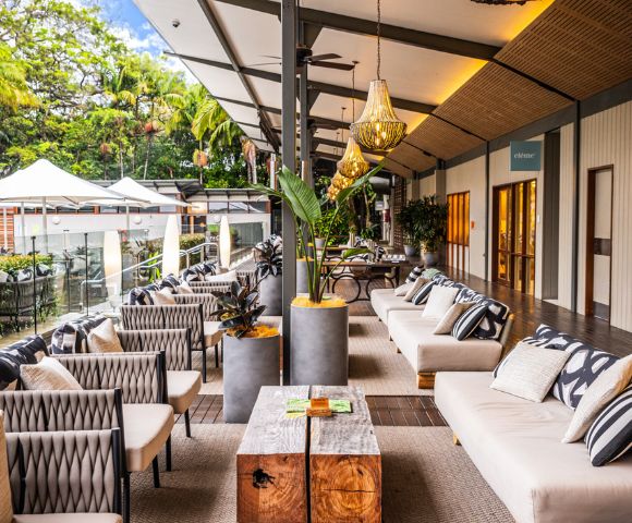 Spacious outdoor patio with plush sofas, patterned cushions, and modern decor. Hanging lights and plants add warmth and elegance, with lush greenery visible.