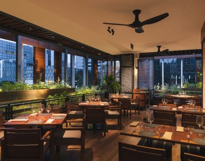 Chic restaurant interior at dusk with wooden tables, neatly set with glasses and napkins. Warm lighting, lush green plants, and large windows create a cozy ambiance.
