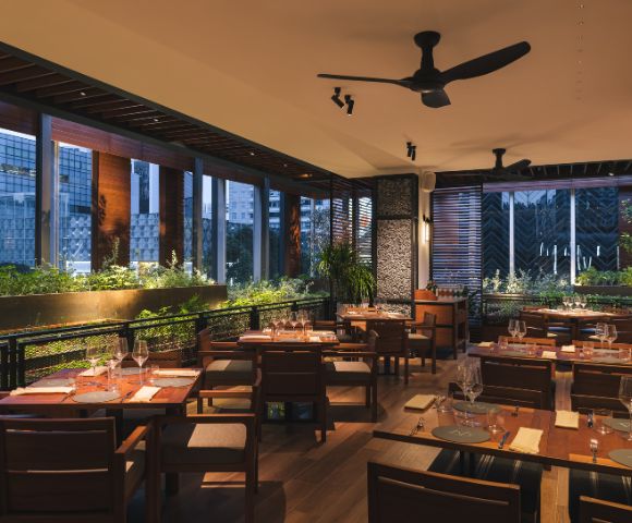 Chic restaurant interior at dusk with wooden tables, neatly set with glasses and napkins. Warm lighting, lush green plants, and large windows create a cozy ambiance.