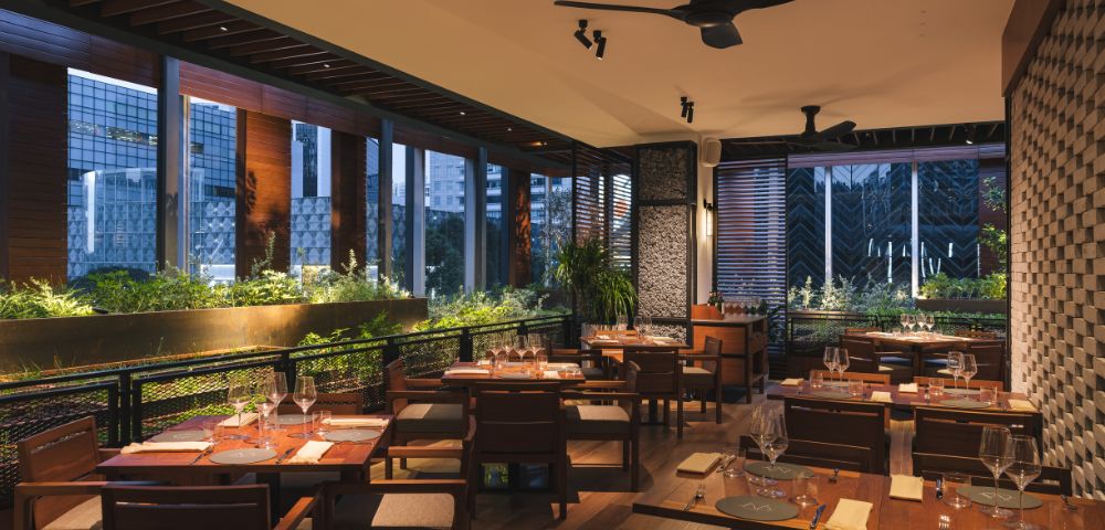 Chic restaurant interior at dusk with wooden tables, neatly set with glasses and napkins. Warm lighting, lush green plants, and large windows create a cozy ambiance.