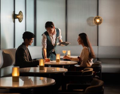 A waiter serves drinks to a man and woman seated at a dimly lit, elegant restaurant table. Warm lighting creates a cozy, intimate atmosphere.