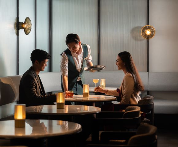 A waiter serves drinks to a man and woman seated at a dimly lit, elegant restaurant table. Warm lighting creates a cozy, intimate atmosphere.