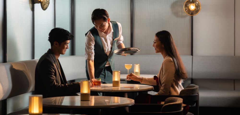 A waiter serves drinks to a man and woman seated at a dimly lit, elegant restaurant table. Warm lighting creates a cozy, intimate atmosphere.