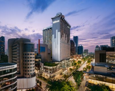 A tall, modern hotel building with the Hilton logo is illuminated at dusk. Surrounded by cityscape and trees, the scene exudes a vibrant urban energy.