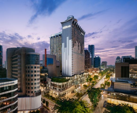 A tall, modern hotel building with the Hilton logo is illuminated at dusk. Surrounded by cityscape and trees, the scene exudes a vibrant urban energy.
