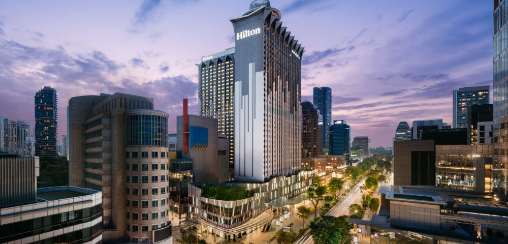 A tall, modern hotel building with the Hilton logo is illuminated at dusk. Surrounded by cityscape and trees, the scene exudes a vibrant urban energy.