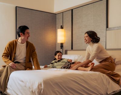 A family of three enjoys a joyful moment on a bed in a cozy room. Parents sit at the edge, smiling at their child who laughs playfully in the center.