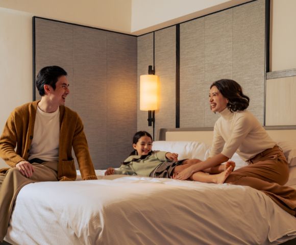 A family of three enjoys a joyful moment on a bed in a cozy room. Parents sit at the edge, smiling at their child who laughs playfully in the center.