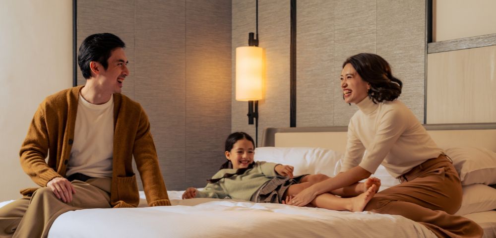 A family of three enjoys a joyful moment on a bed in a cozy room. Parents sit at the edge, smiling at their child who laughs playfully in the center.