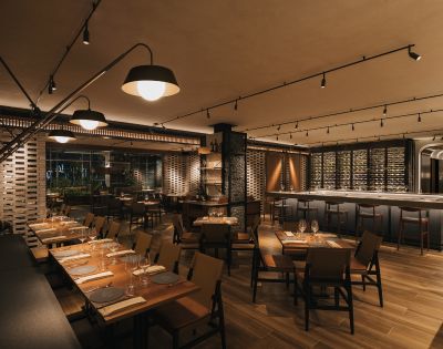 Upscale restaurant interior with warm lighting, wooden floors, and modern furnishings. Tables are set for dining, and a well-stocked bar is visible. Cozy ambiance.