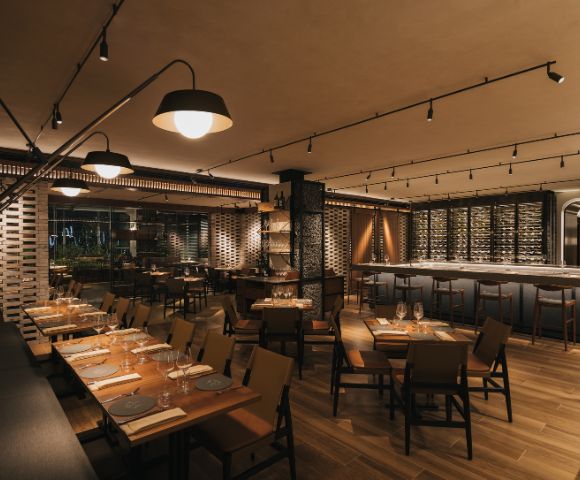 Upscale restaurant interior with warm lighting, wooden floors, and modern furnishings. Tables are set for dining, and a well-stocked bar is visible. Cozy ambiance.