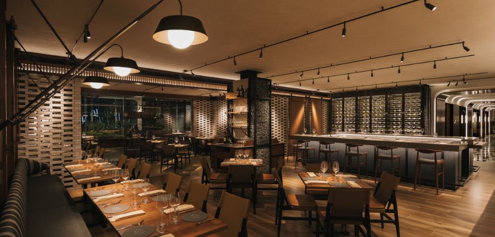Upscale restaurant interior with warm lighting, wooden floors, and modern furnishings. Tables are set for dining, and a well-stocked bar is visible. Cozy ambiance.