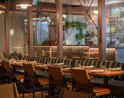 Modern restaurant interior with a long wooden table, cushioned chairs, and neatly set plates. Warm lighting and glass wall reveal a bar, conveying a cozy atmosphere.