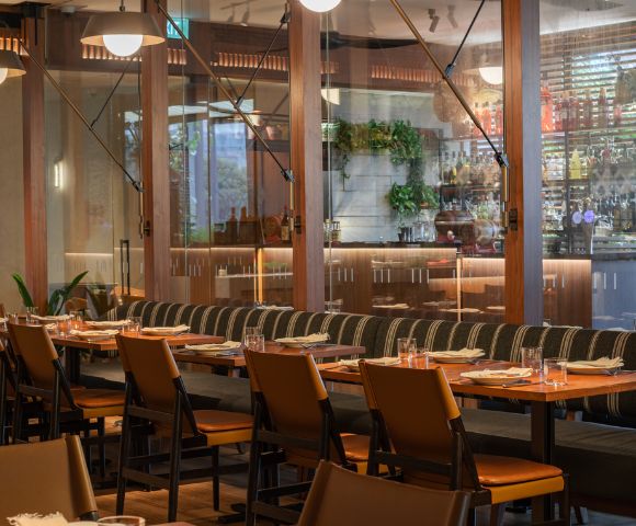 Modern restaurant interior with a long wooden table, cushioned chairs, and neatly set plates. Warm lighting and glass wall reveal a bar, conveying a cozy atmosphere.