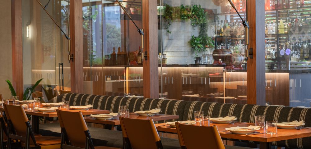 Modern restaurant interior with a long wooden table, cushioned chairs, and neatly set plates. Warm lighting and glass wall reveal a bar, conveying a cozy atmosphere.