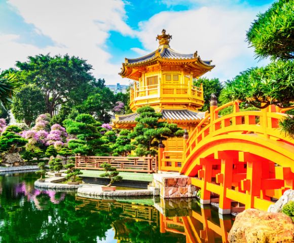 Bright yellow pagoda with gray roof in tranquil garden setting. Arched red bridge spans reflective pond; lush greenery and pink blooms surround. Calm atmosphere.
