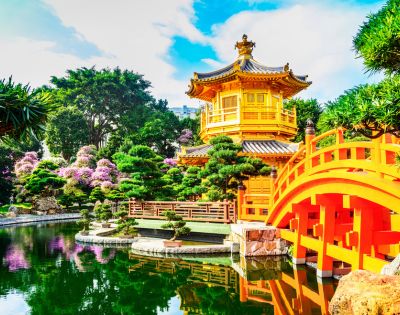 Bright yellow pagoda with gray roof in tranquil garden setting. Arched red bridge spans reflective pond; lush greenery and pink blooms surround. Calm atmosphere.