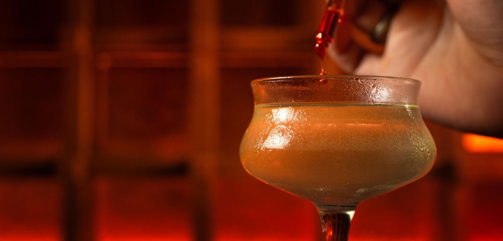 A close-up of a hand using a dropper to add liquid to a frosted cocktail glass. The background is warmly lit, creating a cozy, intimate atmosphere.