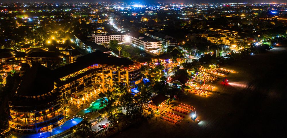 A vibrant night aerial view of a coastal city with illuminated buildings, including hotels by the beach, casting a warm, inviting glow.