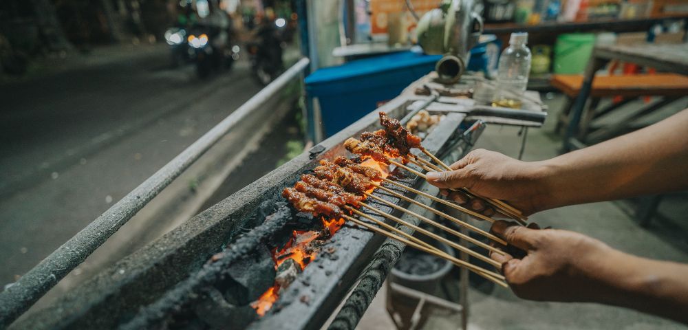 Hands grill meat skewers over glowing coals on a street food stall at night, evoking a warm, bustling atmosphere with rich, savory aromas.