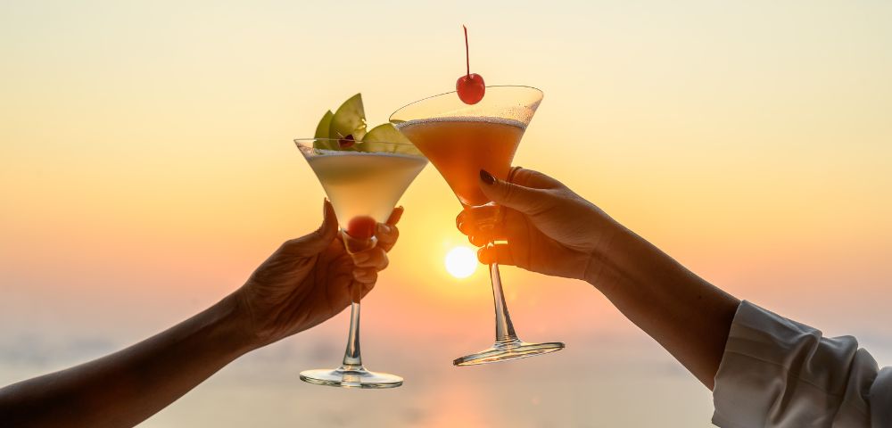 Two hands raise cocktail glasses, one with a cherry garnish, the other with apple slices, against a warm, glowing sunset, conveying cheer and celebration.