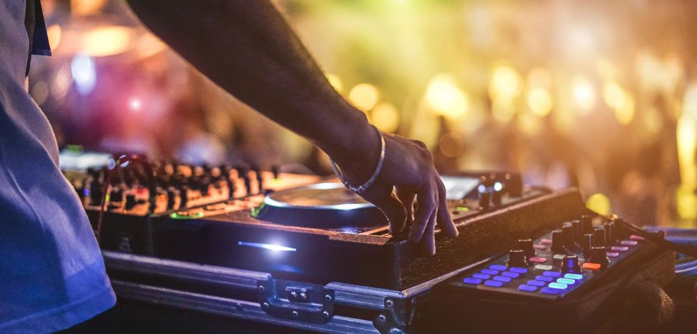 A DJ operates a mixing console, with colorful buttons and dials glowing. Blurred lights and a crowd in the background create an energetic, vibrant atmosphere.