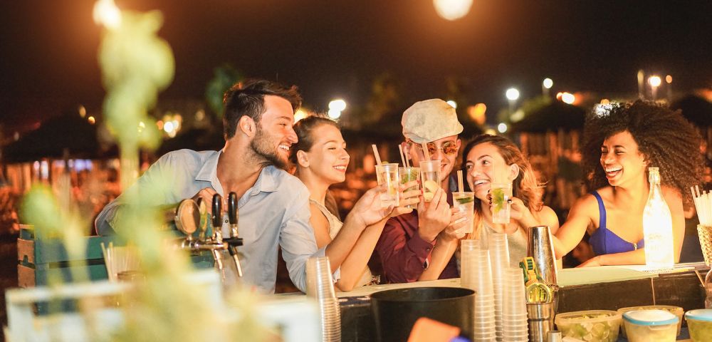 A group of five friends enjoying drinks at a lively beach bar at night. They're laughing and toasting with cocktails, surrounded by warm lighting.