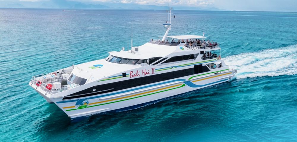 A sleek white ferry with colorful stripes sails on clear blue ocean waters under a partially cloudy sky, conveying a sense of serenity and adventure.