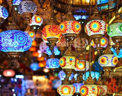 Colorful mosaic lamps hang in a market, emitting warm, vibrant hues of blue, red, and gold. The atmosphere is lively and rich with cultural charm.