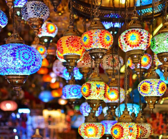 Colorful mosaic lamps hang in a market, emitting warm, vibrant hues of blue, red, and gold. The atmosphere is lively and rich with cultural charm.