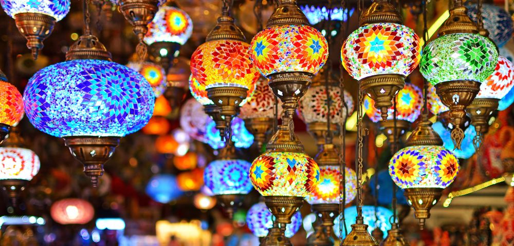 Colorful mosaic lamps hang in a market, emitting warm, vibrant hues of blue, red, and gold. The atmosphere is lively and rich with cultural charm.