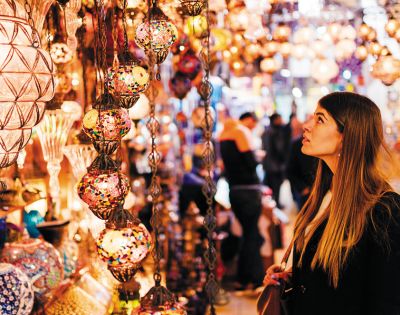 A woman gazes at colorful, intricately designed hanging lamps in a vibrant market. The scene is bustling, with a warm, lively atmosphere.