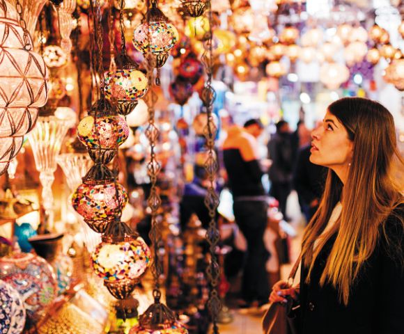 A woman gazes at colorful, intricately designed hanging lamps in a vibrant market. The scene is bustling, with a warm, lively atmosphere.