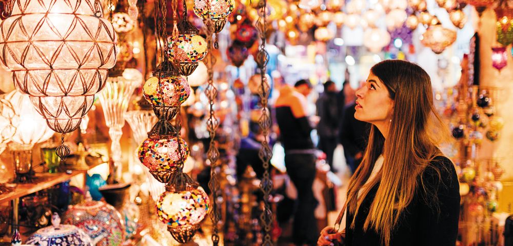 A woman gazes at colorful, intricately designed hanging lamps in a vibrant market. The scene is bustling, with a warm, lively atmosphere.