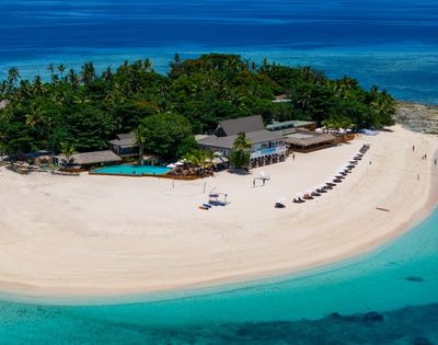 Aerial view of a tropical island with lush greenery, sandy beach, and crystal-clear blue water. Beach chairs are aligned along the shore beside a resort.