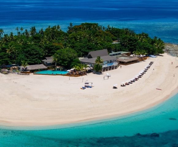 Aerial view of a tropical island with lush greenery, sandy beach, and crystal-clear blue water. Beach chairs are aligned along the shore beside a resort.