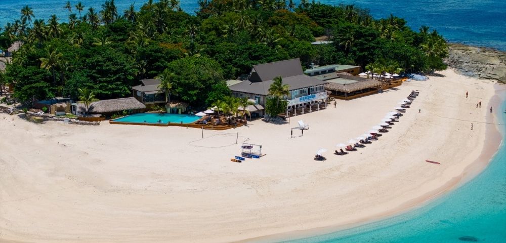 Aerial view of a tropical island with lush greenery, sandy beach, and crystal-clear blue water. Beach chairs are aligned along the shore beside a resort.