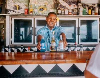 Smiling bartender in a tropical shirt serves colorful drinks at a vibrant bar with decorative tilework, creating a lively, welcoming atmosphere.