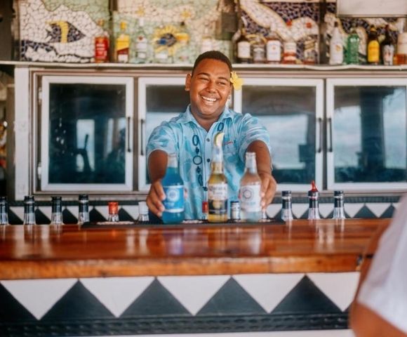 Smiling bartender in a tropical shirt serves colorful drinks at a vibrant bar with decorative tilework, creating a lively, welcoming atmosphere.