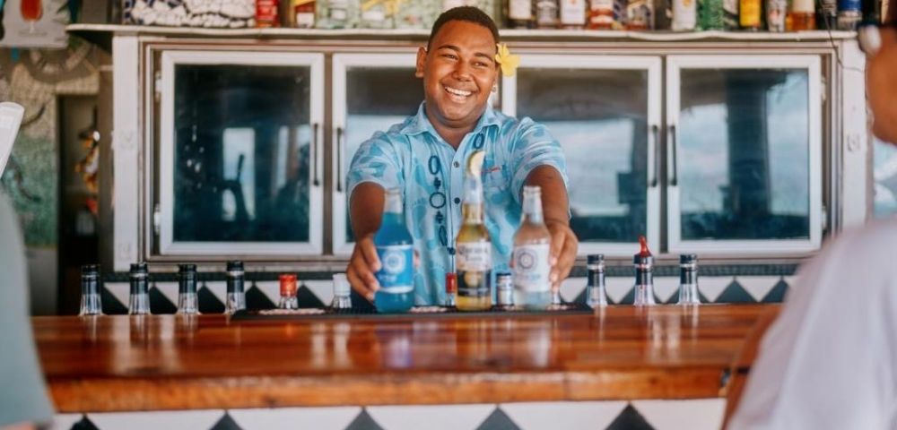 Smiling bartender in a tropical shirt serves colorful drinks at a vibrant bar with decorative tilework, creating a lively, welcoming atmosphere.