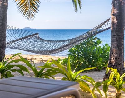 A hammock hangs between palm trees on a tropical beach, with vibrant green plants and a serene ocean view, conveying a tranquil, relaxing vibe.