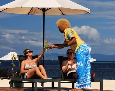 Couple lounging on beach chairs under an umbrella, receiving colorful cocktails from a server in bright attire, with a scenic ocean backdrop. Relaxed vibe.