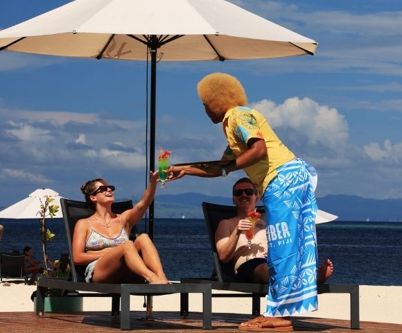 Couple lounging on beach chairs under an umbrella, receiving colorful cocktails from a server in bright attire, with a scenic ocean backdrop. Relaxed vibe.