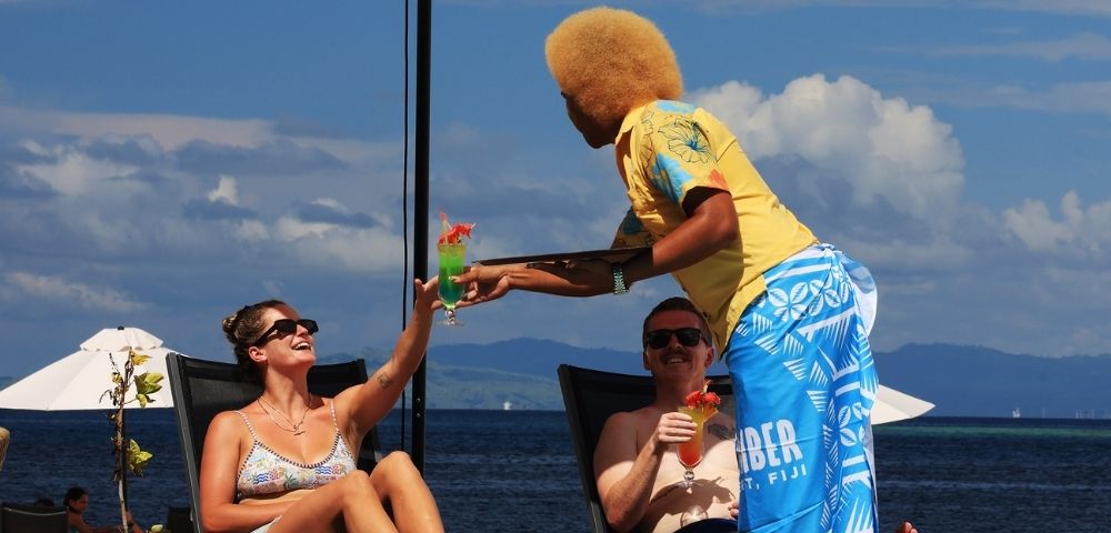 Couple lounging on beach chairs under an umbrella, receiving colorful cocktails from a server in bright attire, with a scenic ocean backdrop. Relaxed vibe.